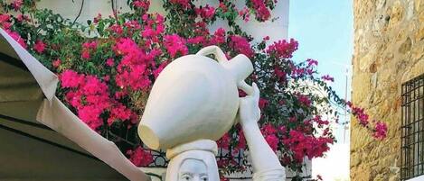 Une statue d’une femme tenant une cruche, entourée de plantes en pot et d’un bâtiment à la façade en pierre.