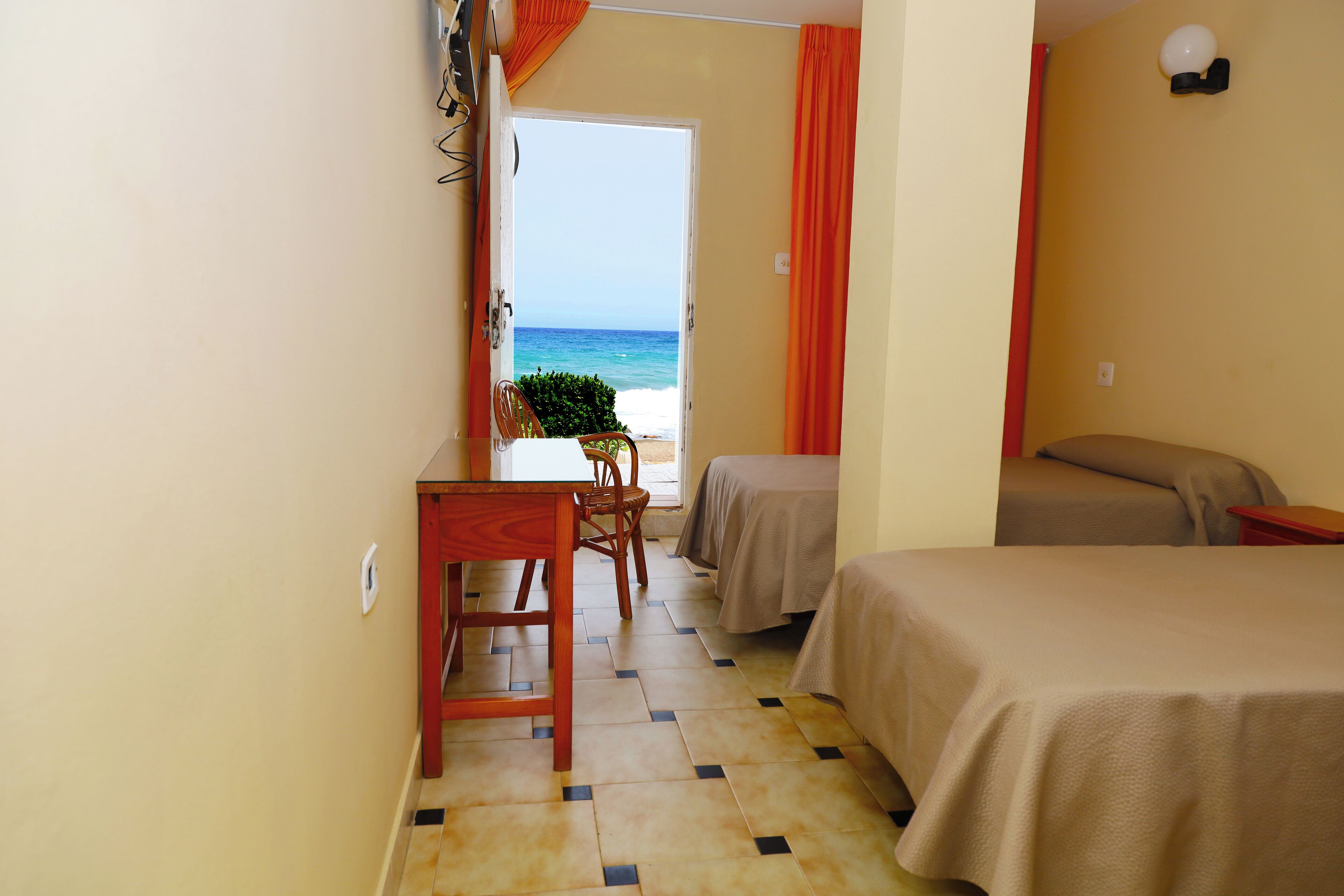 A room with two beds, a desk, and a chair, with a view of the beach and ocean through an open door.