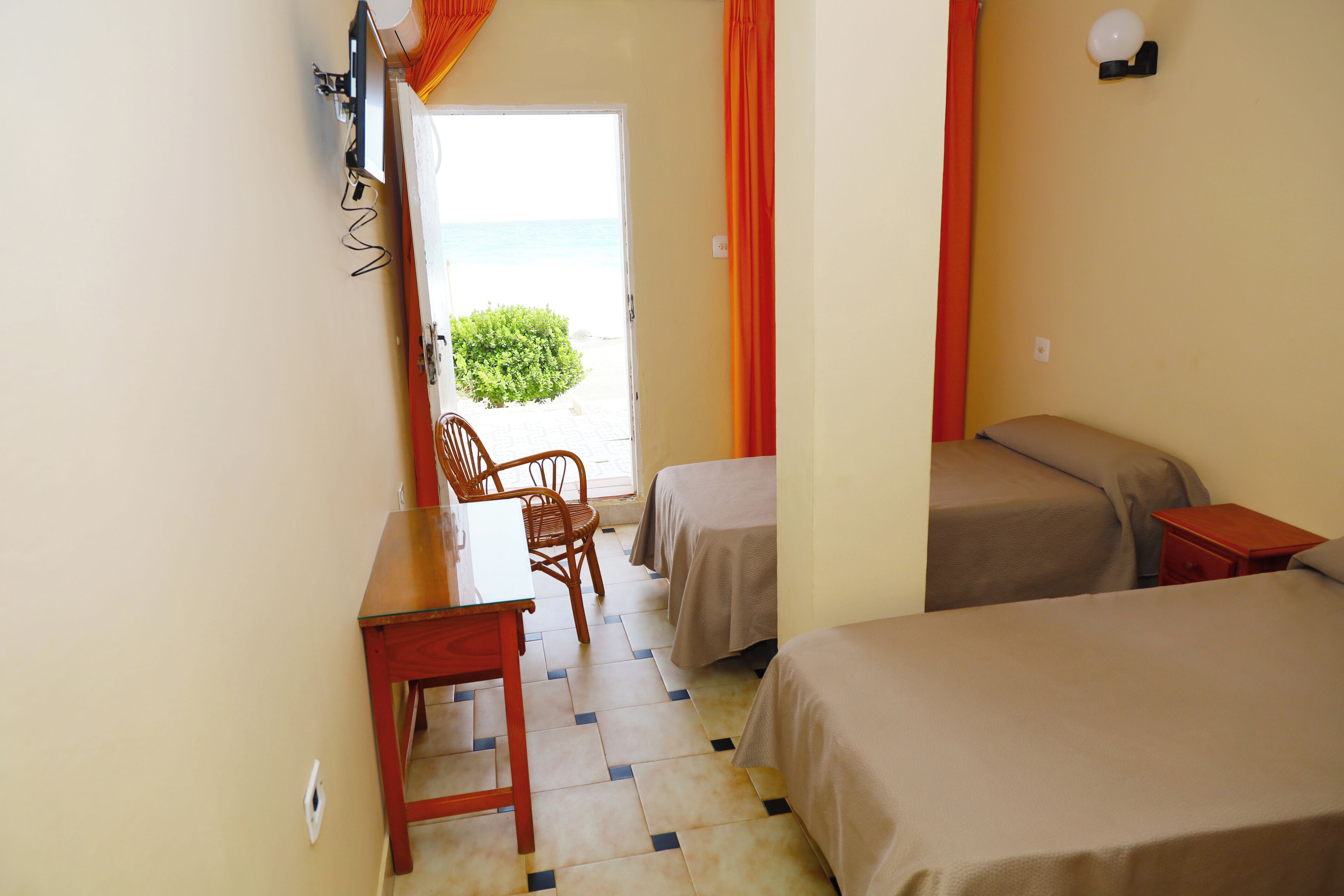Habitación de hotel con dos camas, un escritorio y una silla. Se ve la playa a través de una puerta abierta.