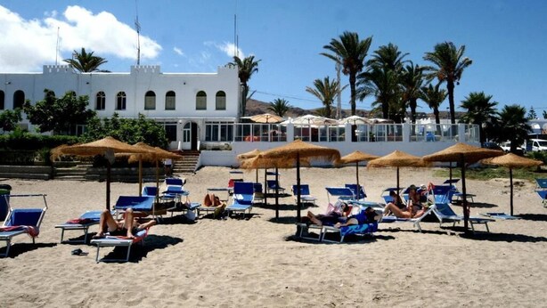 A beach with sun loungers and umbrellas, a white building with arched windows, and palm trees in the background.