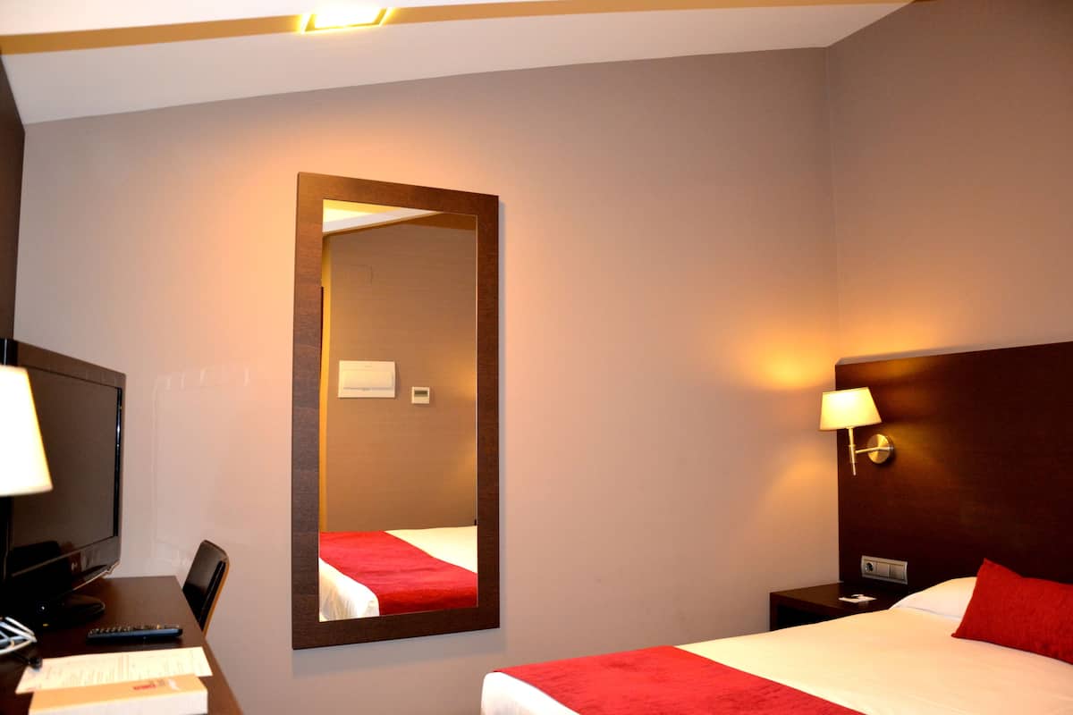 A hotel room with a bed, a desk with a computer, a chair, a nightstand with a lamp, and a large mirror on the wall.