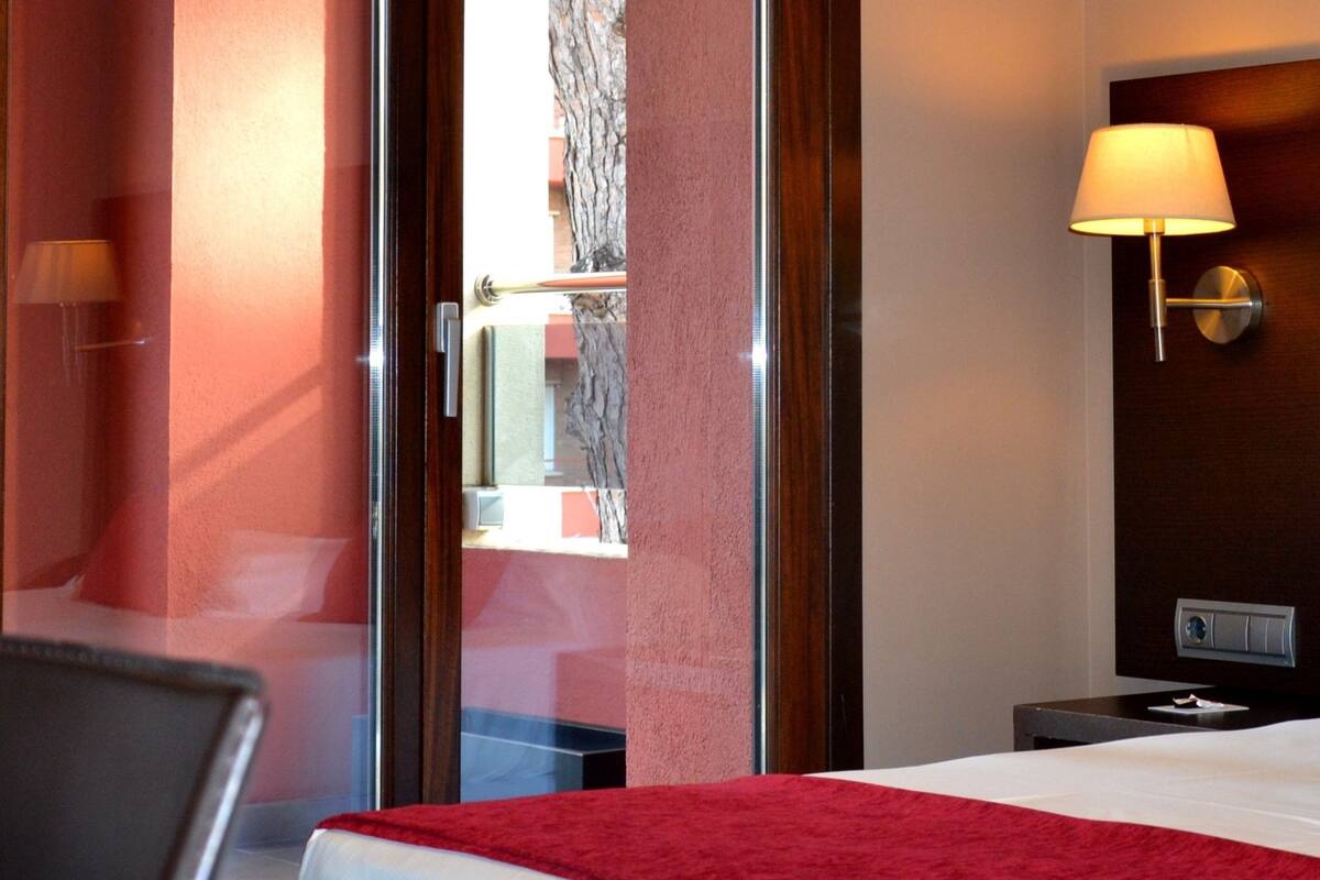 A hotel room with a bed, a nightstand, a lamp, and a view of the outside.