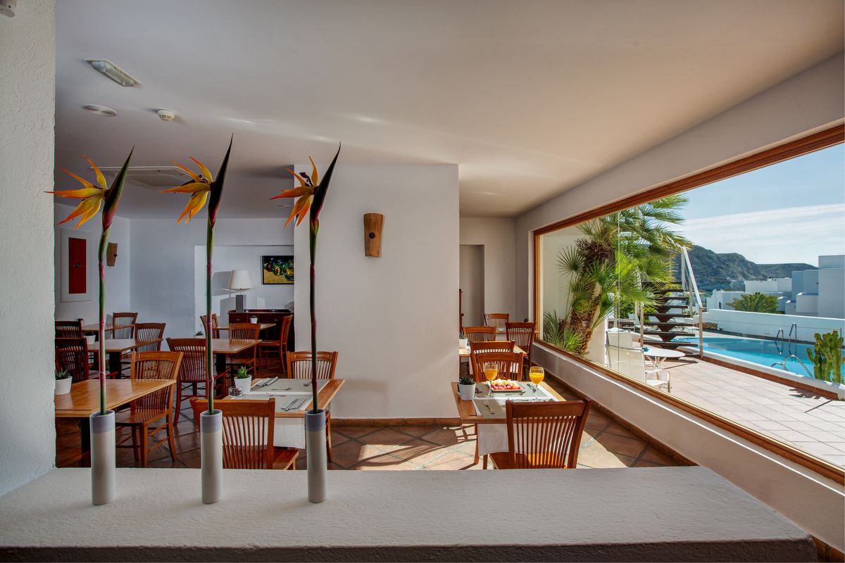 Un comedor moderno con mesas y sillas de madera, un ventanal con vistas a una piscina y montañas.