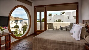 A hotel room with a large bed, a TV, a round window, and a balcony with a view of the sea and white buildings. - Boutique Hotel El Tio Kiko (Nijar)