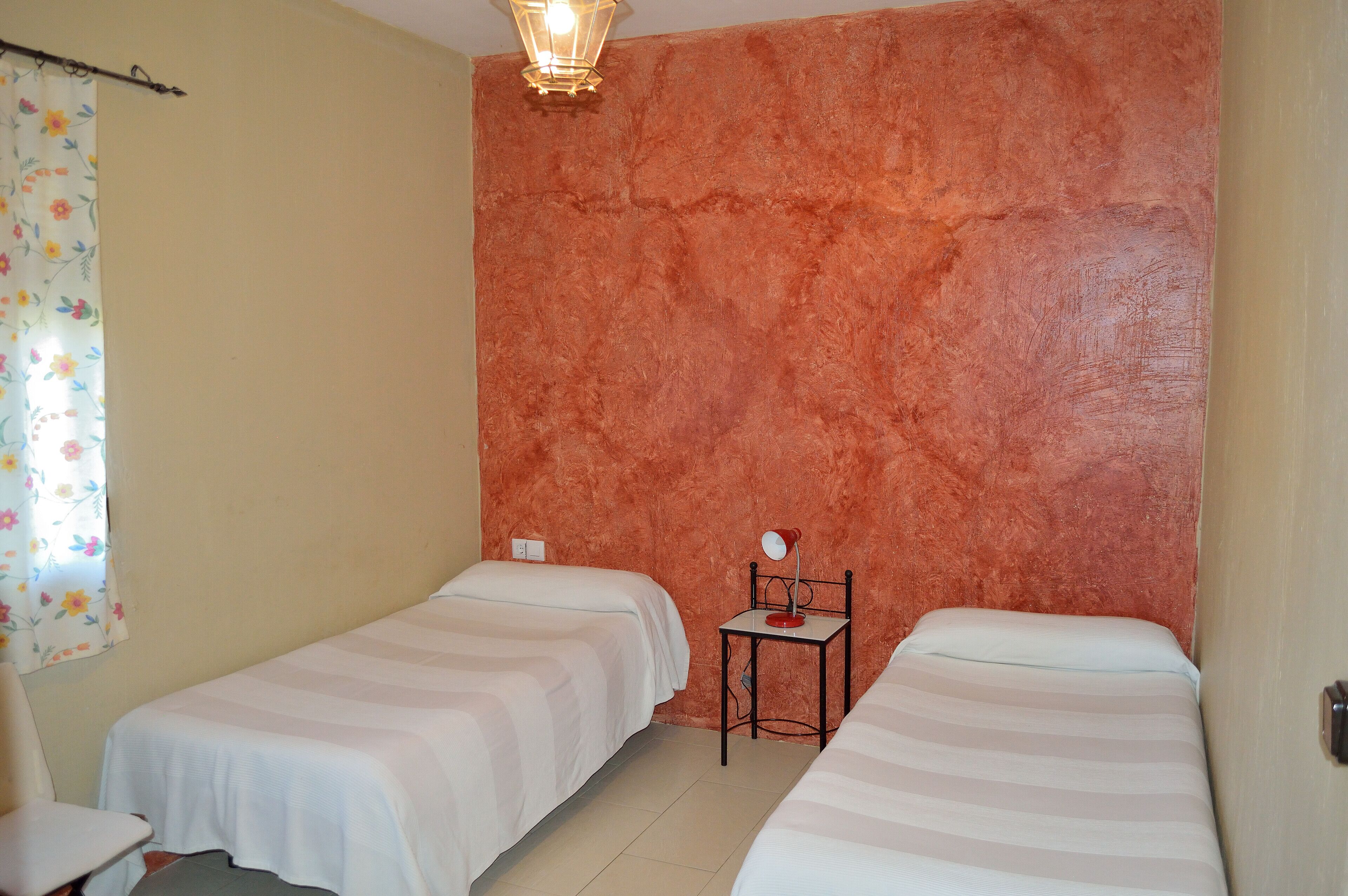 A room with two beds, a small table with a lamp, and a window with a floral curtain.