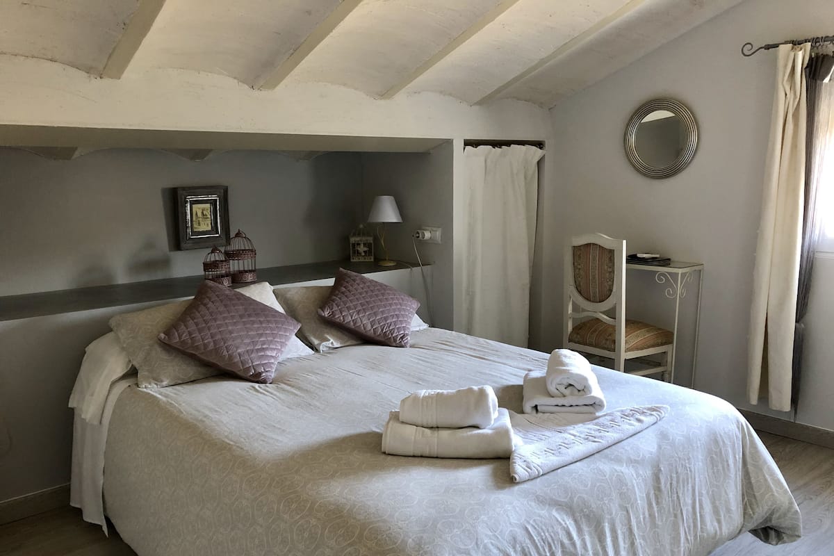 A neatly made bed with pillows, a bedside table with a lamp, a chair, and a mirror on the wall.