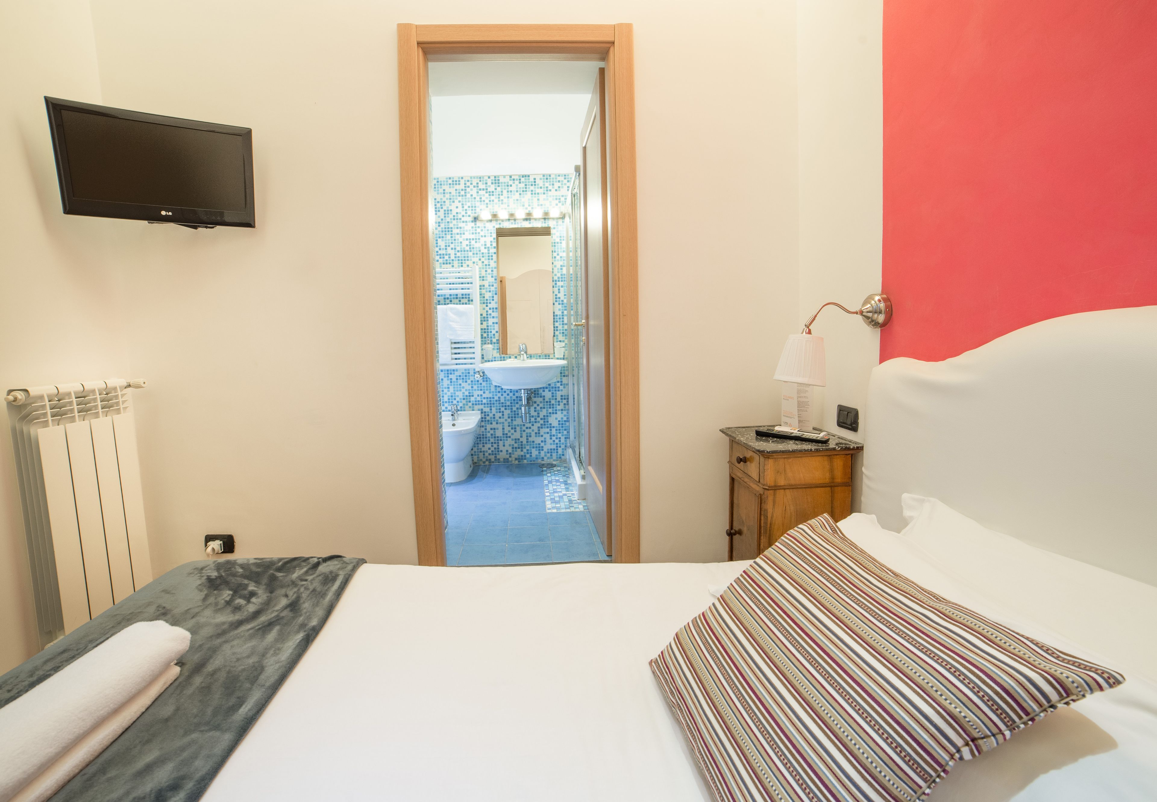 Habitación de hotel con cama, televisión montada en la pared, mesita de noche con lámpara y vista al baño a través de una puerta abierta.
