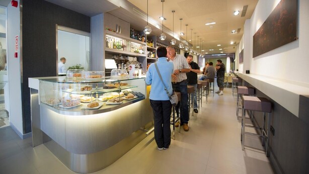 A modern café with a glass display case of pastries, a bar area with stools, and a clean, contemporary interior design. - San Pedro (Langreo)