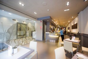 A modern restaurant interior with a glass wall displaying a leaf pattern, a central counter with a person serving, and several tables and chairs. - San Pedro (Langreo)