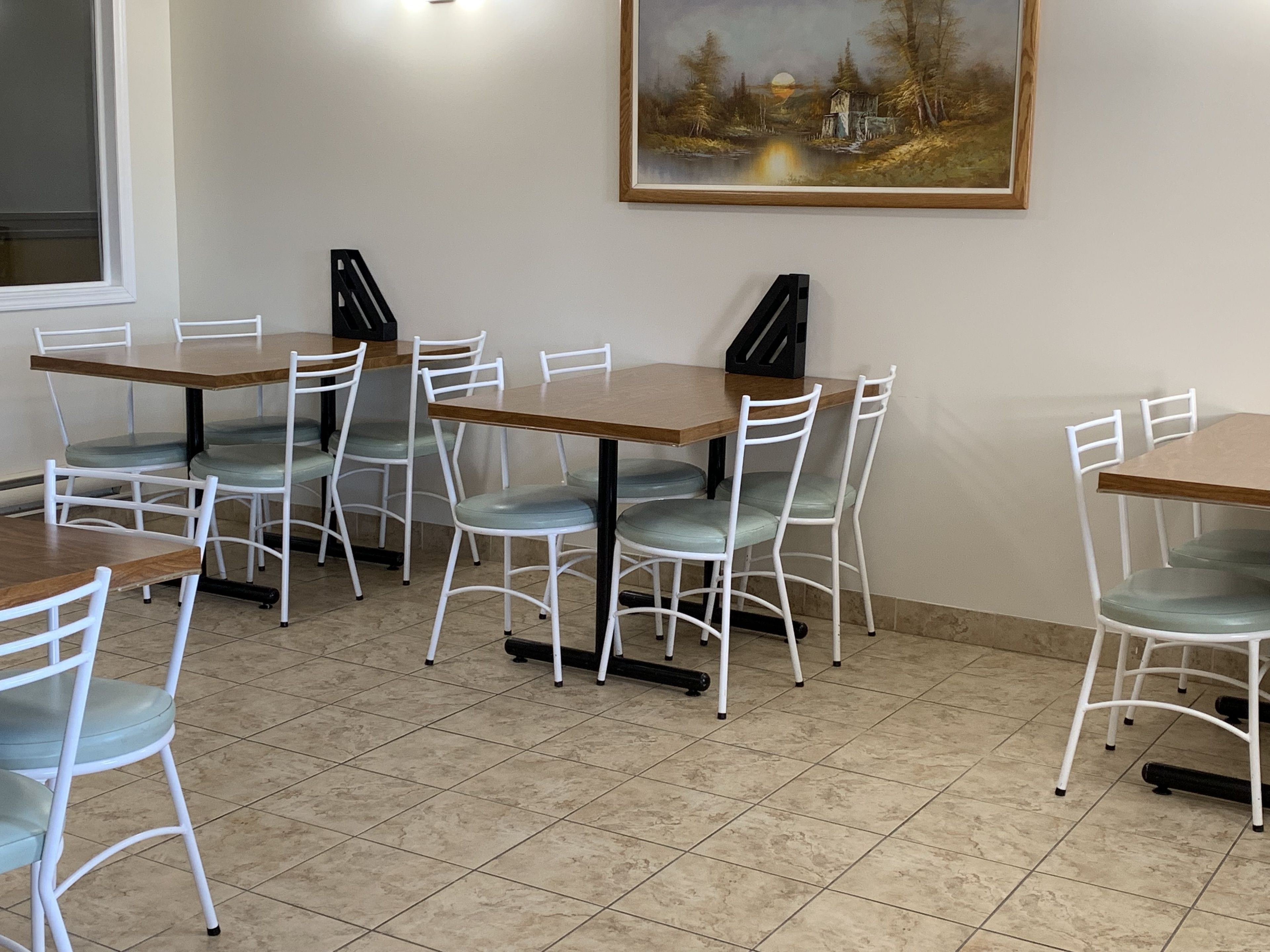 A dining area with six tables and chairs, a painting on the wall, and a window on the left.