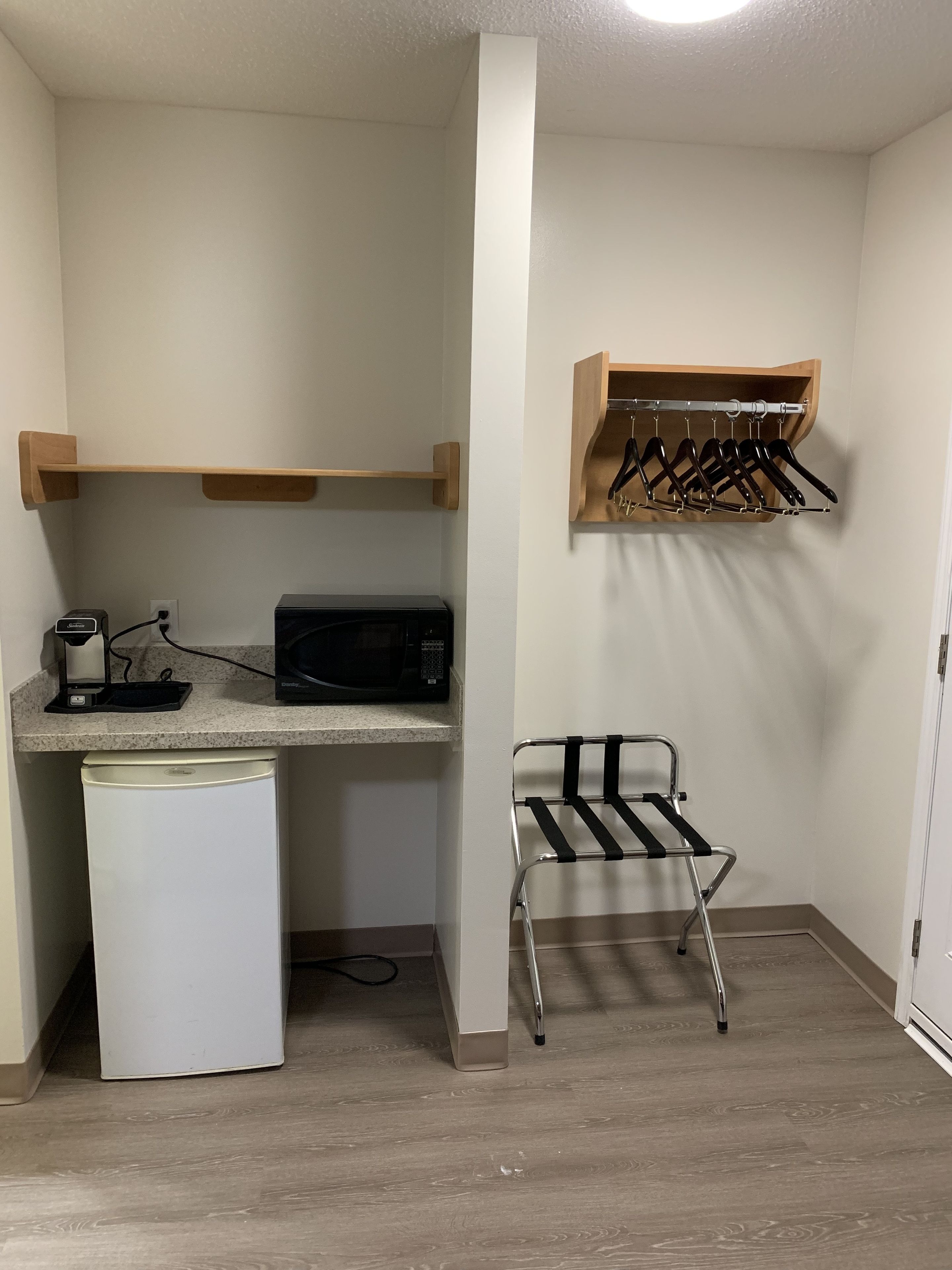 A compact kitchenette with a microwave, coffee maker, and a small refrigerator. There is a metal drying rack and a wooden shelf above it.