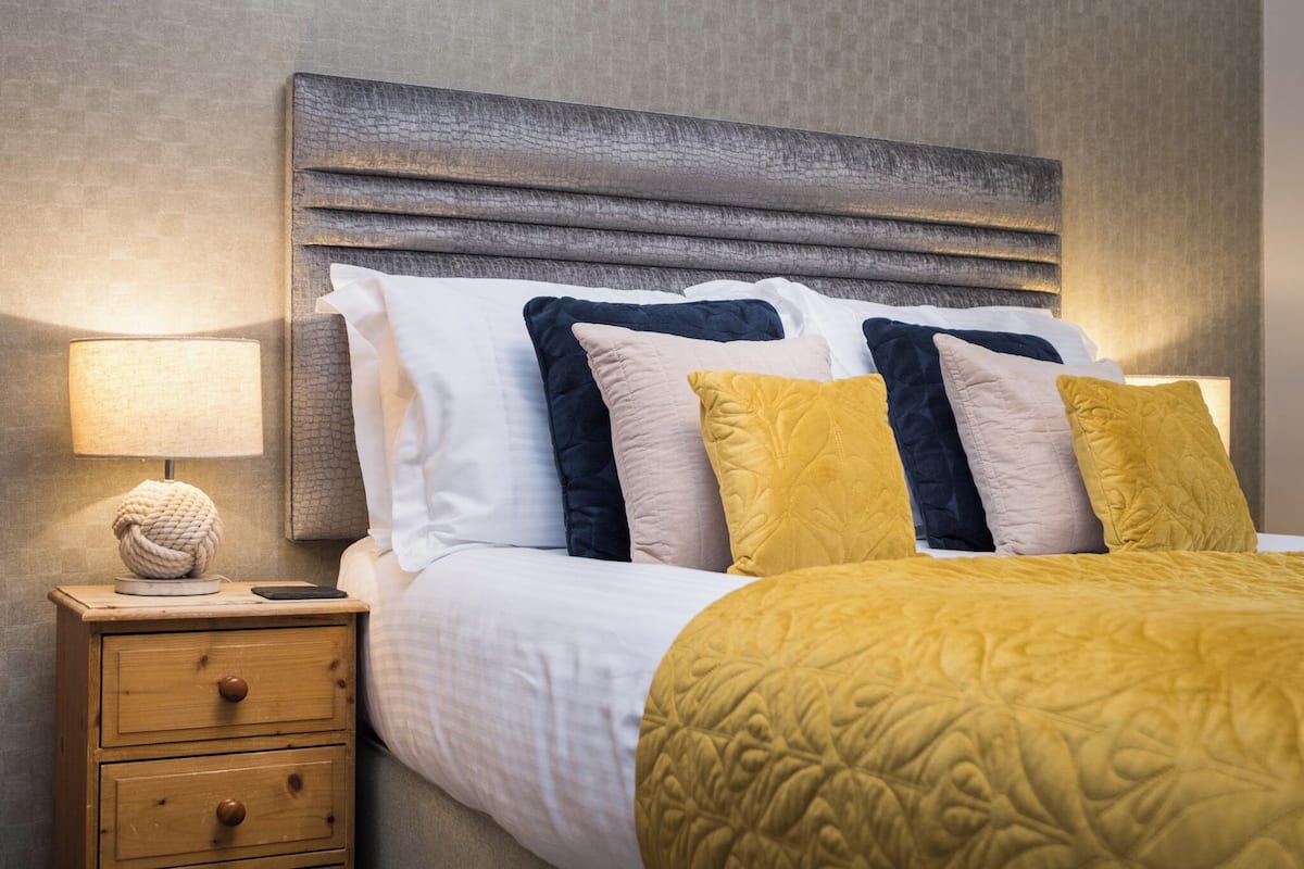 A neatly made bed with a textured yellow bedspread, a wooden nightstand with a lamp and a decorative item, and a textured wall behind the bed.