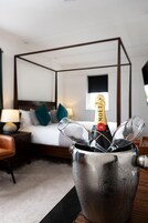 A minibar with a bottle of champagne and glasses, a bed with a wooden headboard, a nightstand with a lamp, and a view of a window.