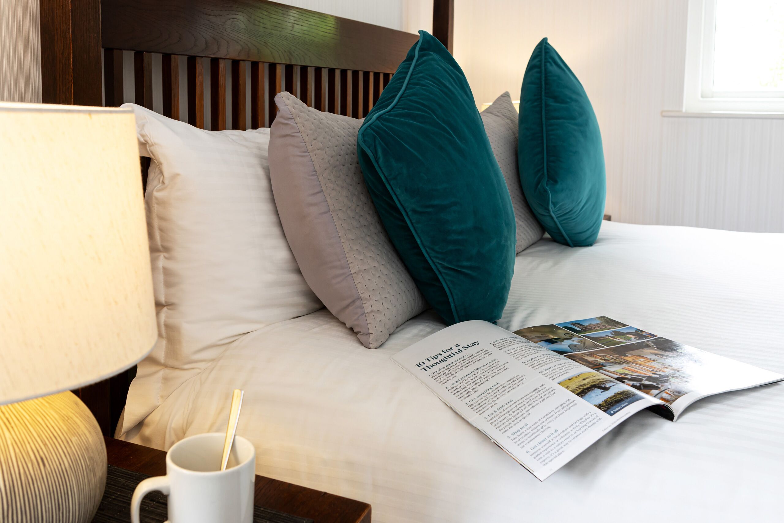 A neatly made bed with a magazine, a white mug, and a lamp on a bedside table.