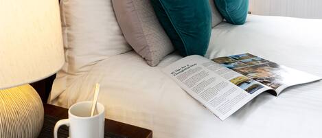 A neatly made bed with a magazine, a white mug, and a lamp on a bedside table.