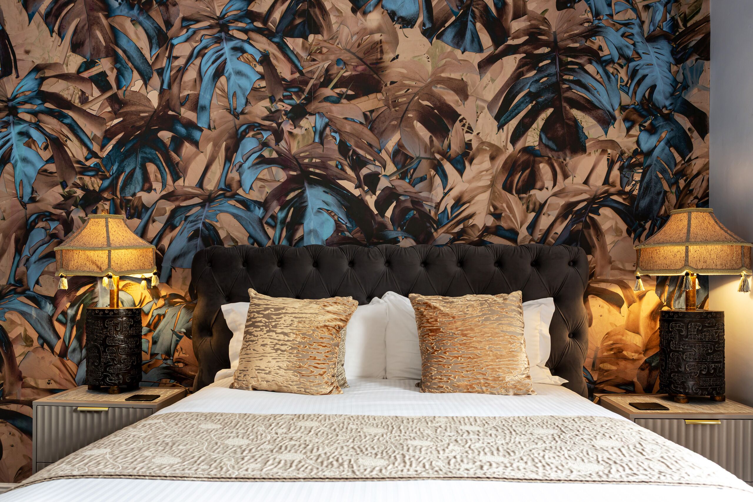 A bedroom with a tufted headboard, two bedside lamps, a patterned bedspread, and a tropical leaf wallpaper.