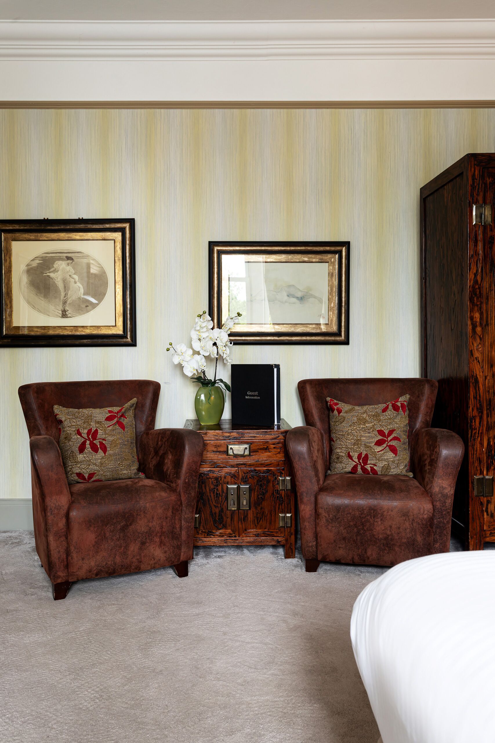 A room with two leather armchairs, a small wooden side table with a vase and a black object, and a large wooden cabinet.