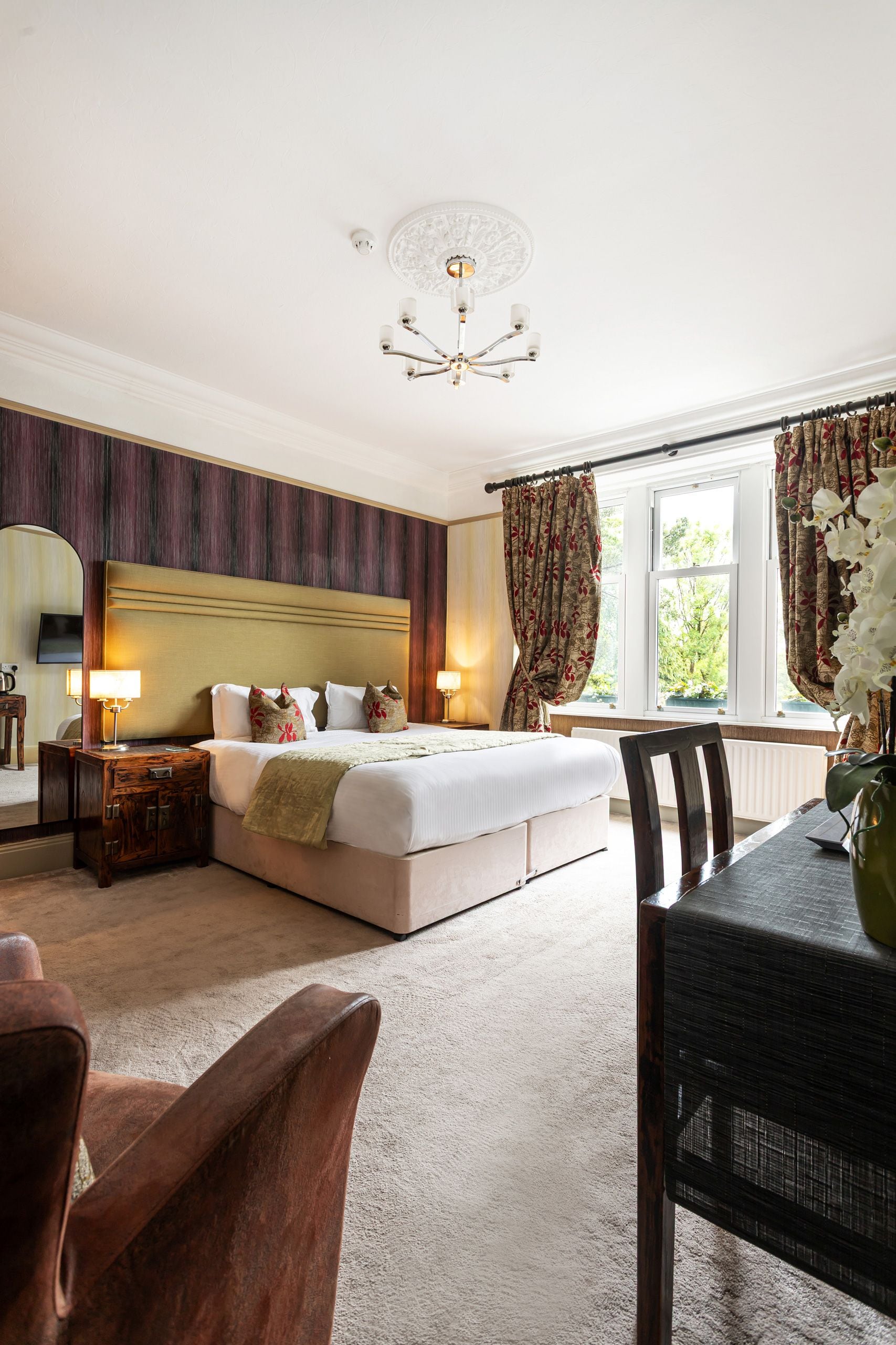 A hotel room with a large bed, a wooden nightstand, a table with a vase, and a view of the outdoors through a window.