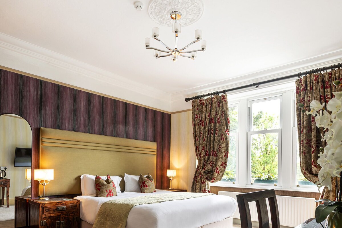 A hotel room with a large bed, a wooden nightstand, a table with a vase, and a view of the outdoors through a window.