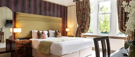 A hotel room with a large bed, a wooden nightstand, a table with a vase, and a view of the outdoors through a window.