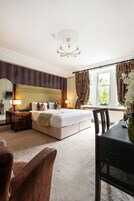 A hotel room with a large bed, a wooden nightstand, a table with a vase, and a view of the outdoors through a window.