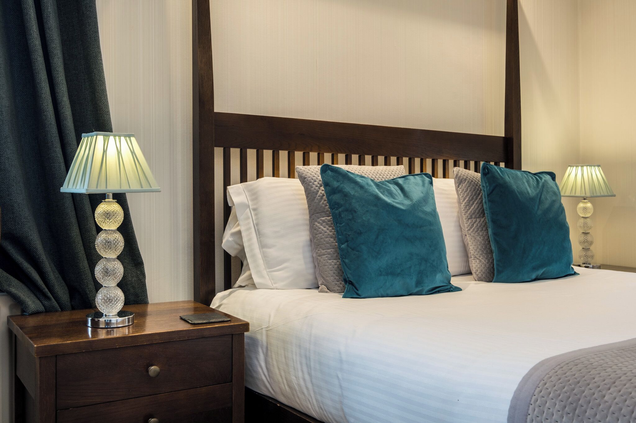 A neatly made bed with white linens and teal pillows, a wooden nightstand with a lamp, and a dark curtain.