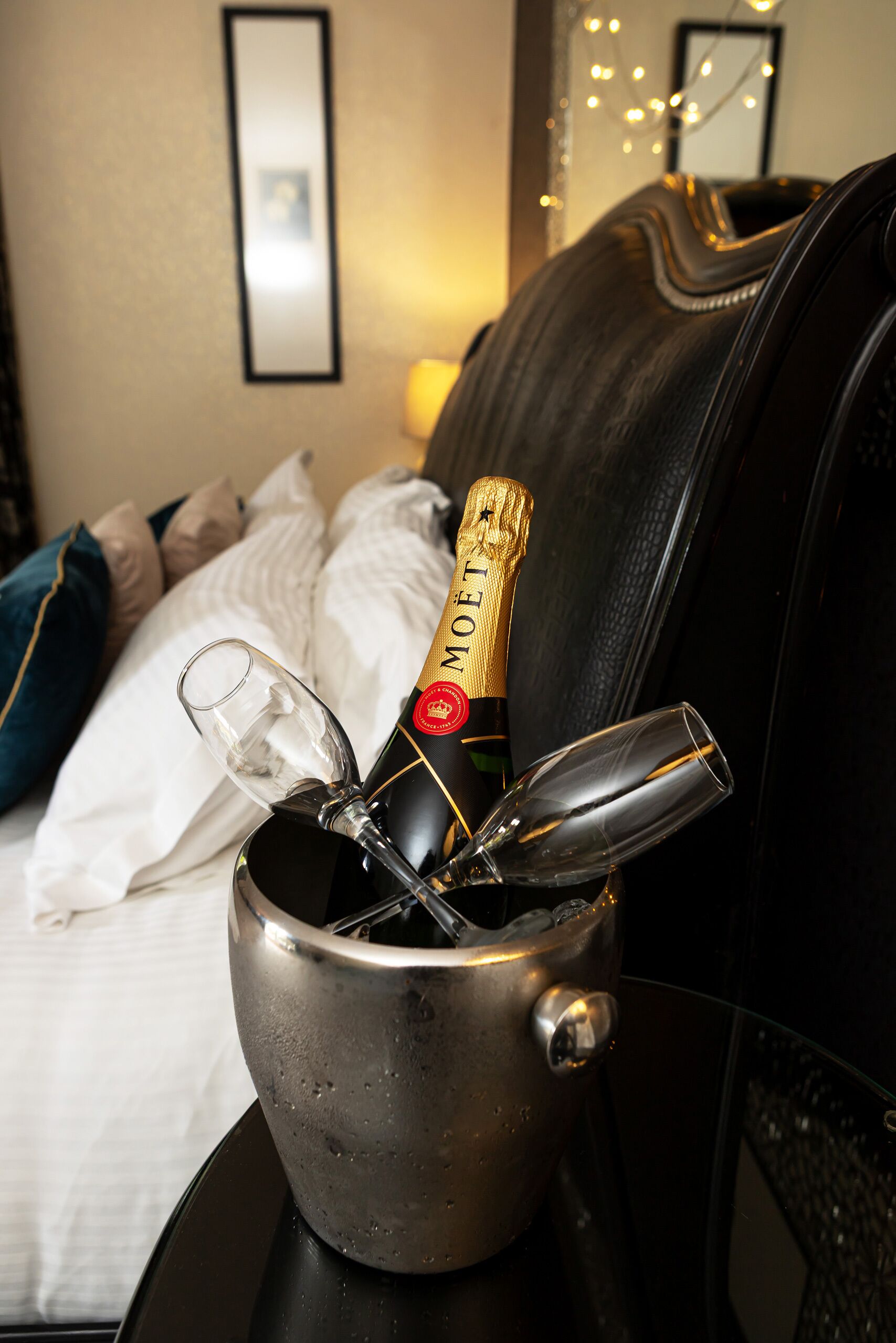 A champagne bottle in a metal bucket on a bed with pillows and a blanket.