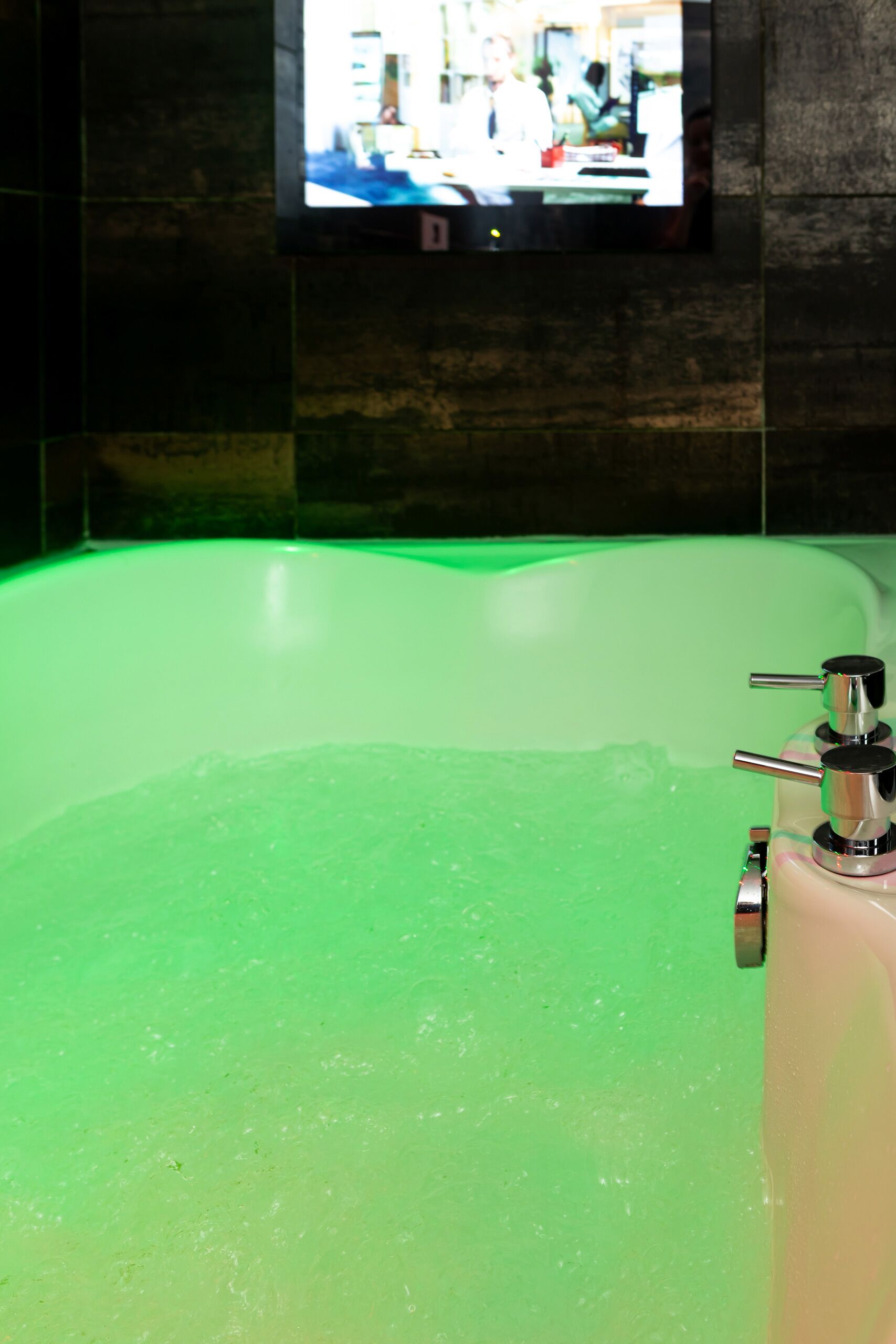 A bathtub with green lighting, a faucet, and a television mounted on the wall.