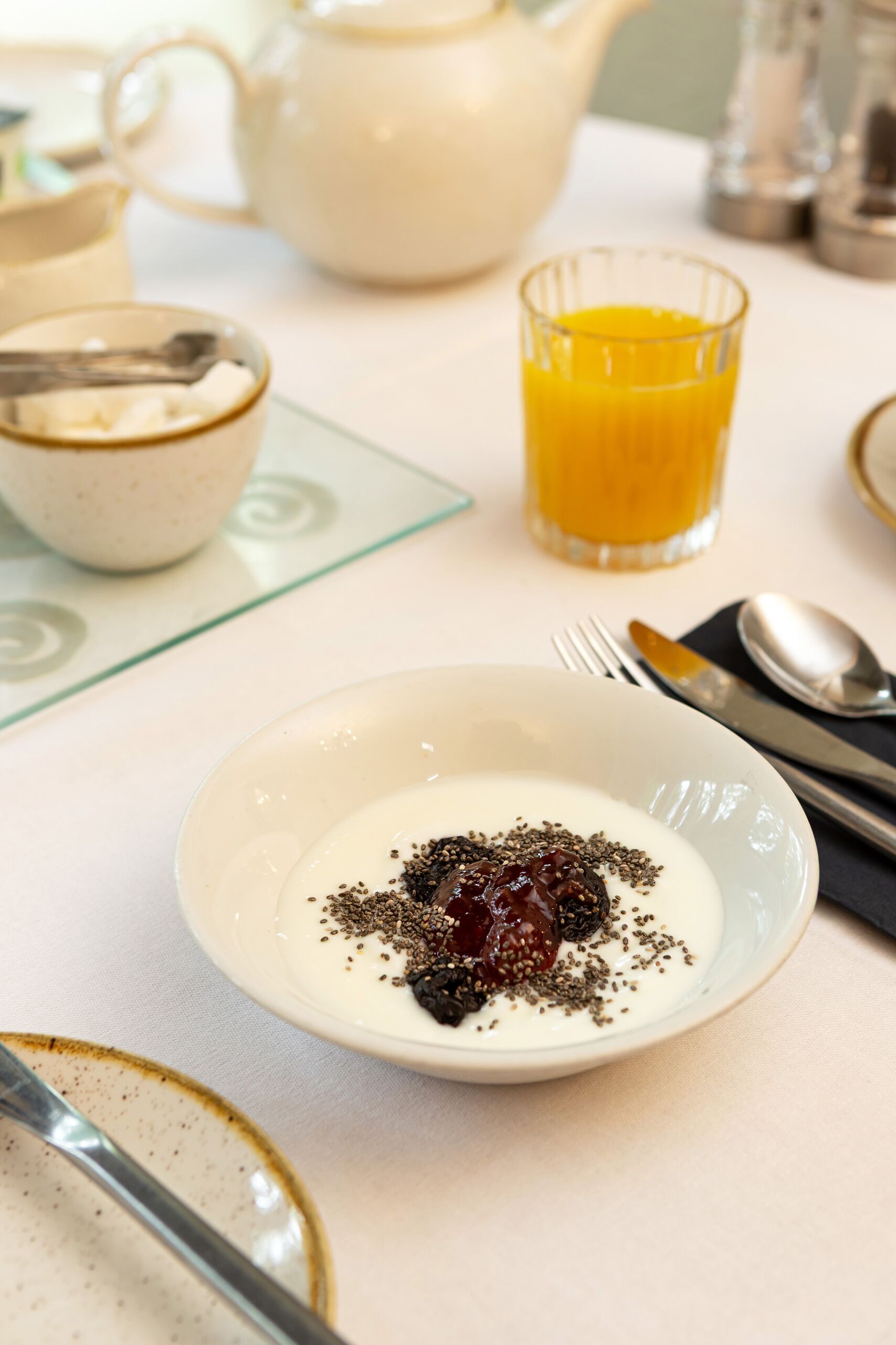 Un desayuno con un plato de yogur cubierto de semillas y una cereza, un vaso de zumo de naranja y una tetera al fondo.
