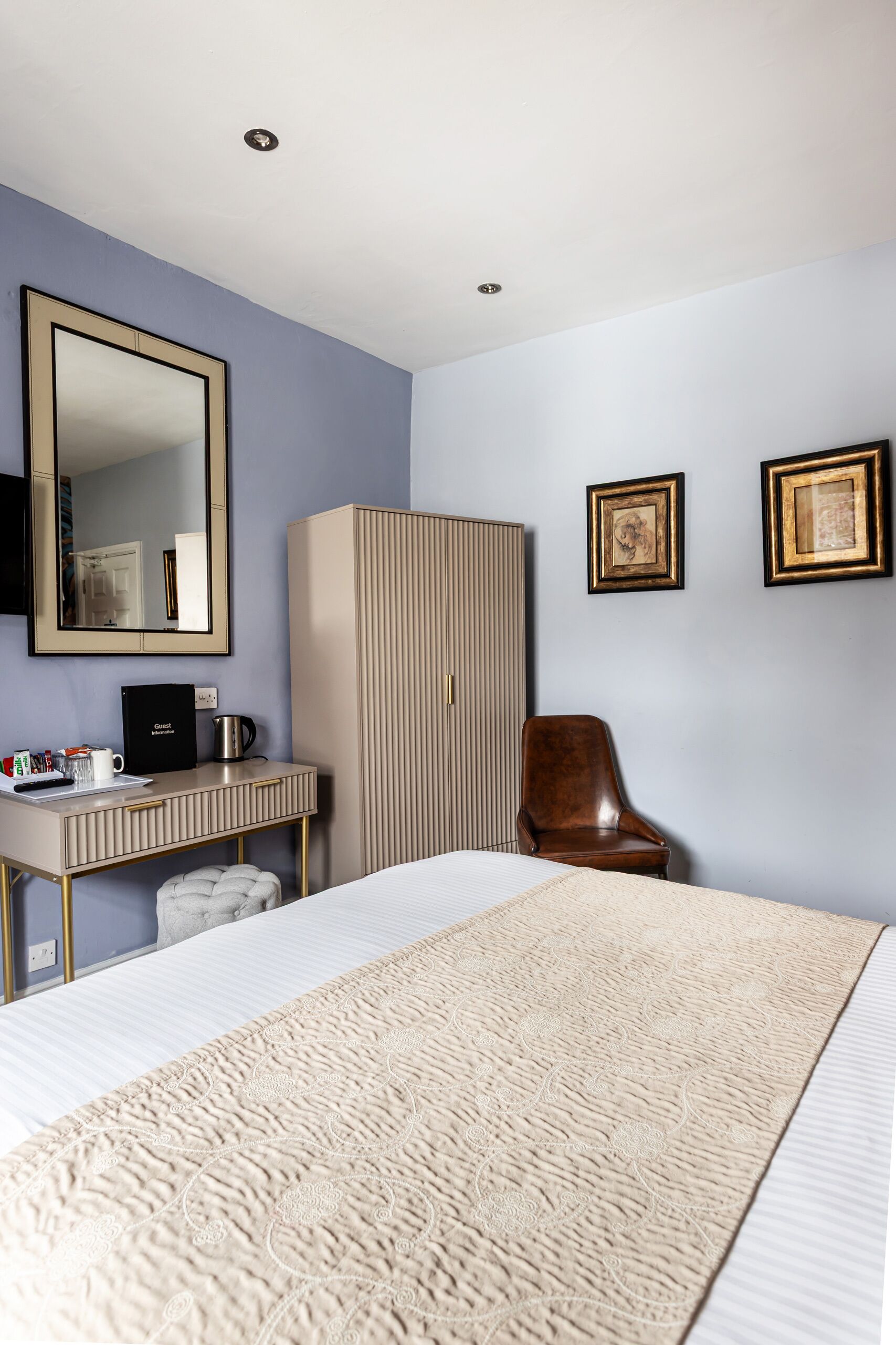 A hotel room with a bed, a desk with a coffee maker, a wardrobe, and two framed pictures on the wall.