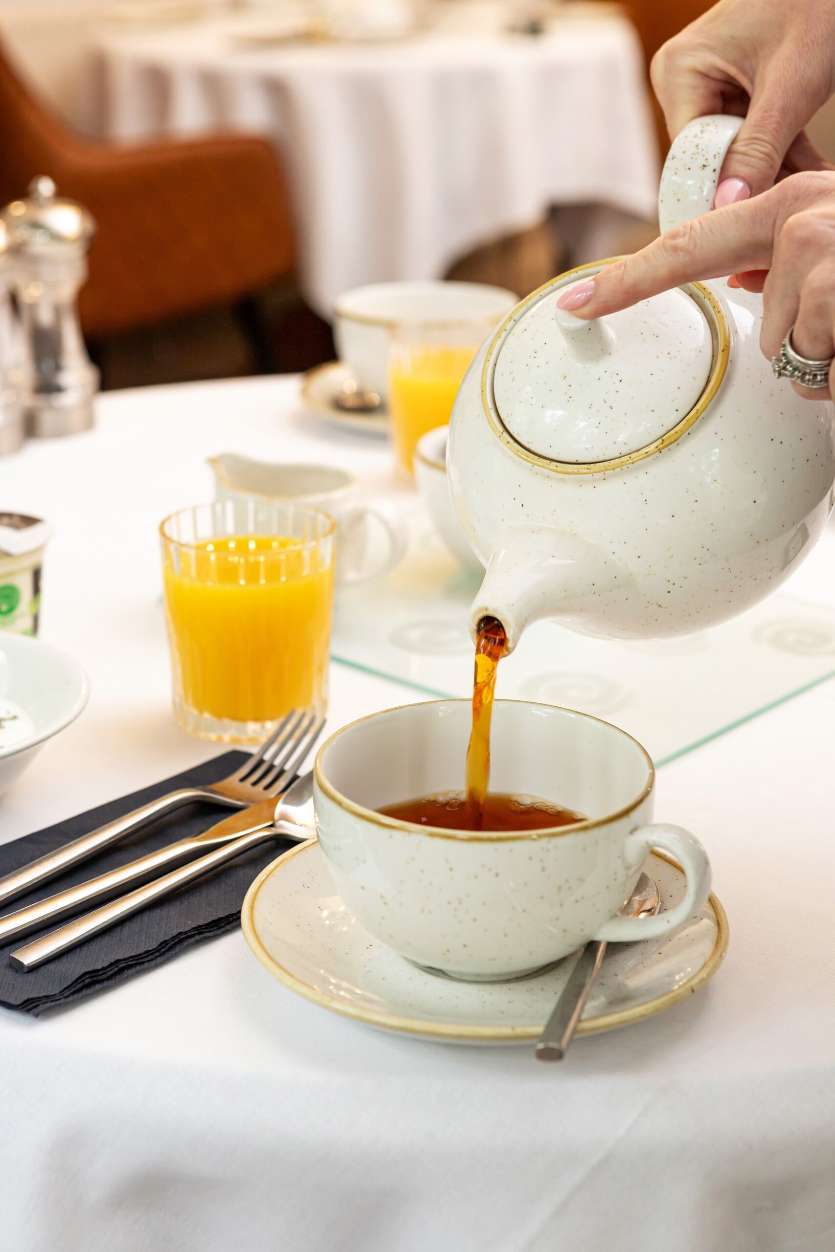 A teapot pouring tea into a cup, a glass of orange juice, and a fork on a table.