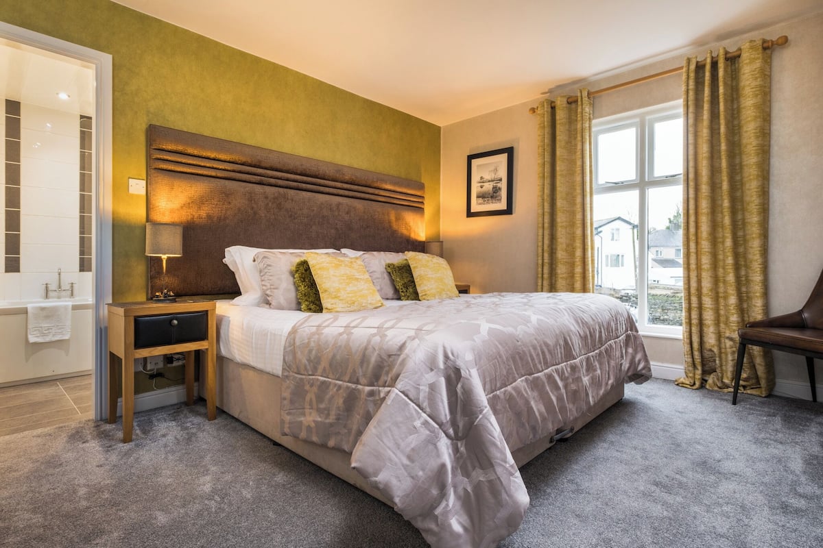 A bedroom with a large bed, a nightstand, a lamp, a chair, and a window with curtains.