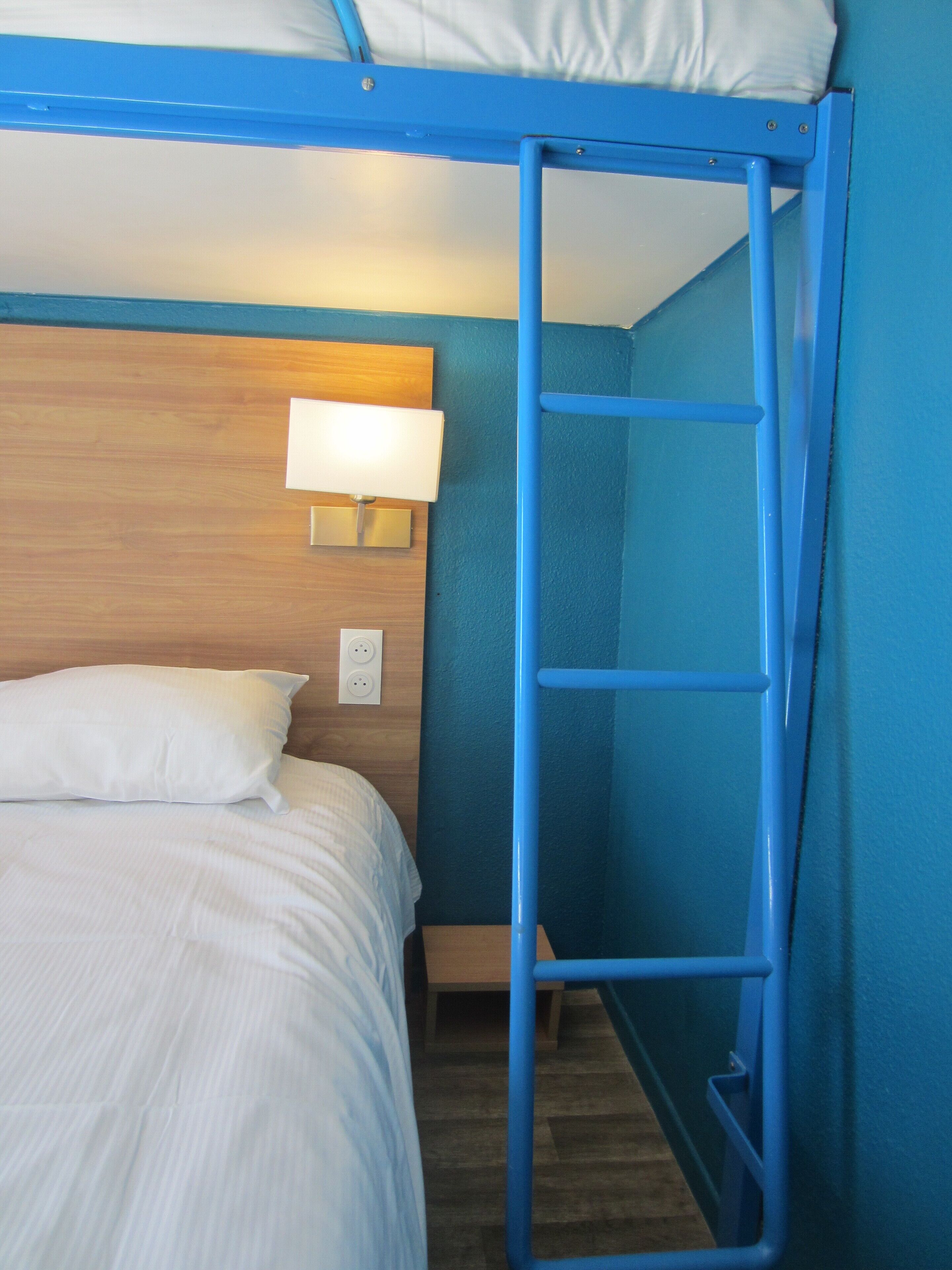Una cama individual superpuesta con escalera, una lámpara de noche y un enchufe en la pared.