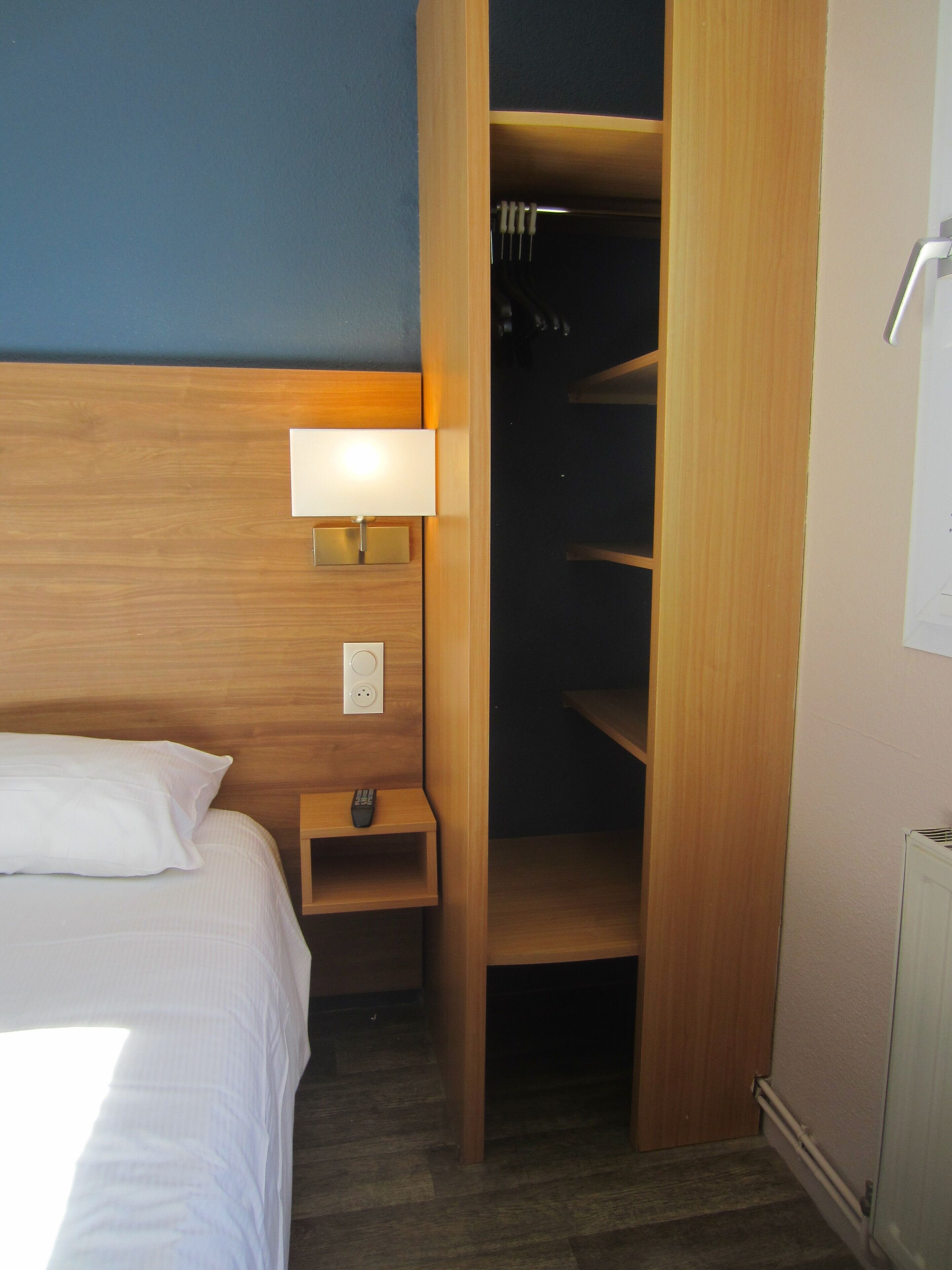 Une chambre d’hôtel avec un lit, une table de chevet avec une lampe, une petite étagère et une armoire en bois avec un miroir.