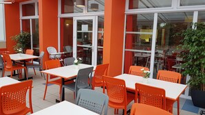 An outdoor seating area with orange and grey chairs and white tables, surrounded by glass windows and a red wall. - The Originals Access, Hôtel les Iris (Berck-sur-Mer)
