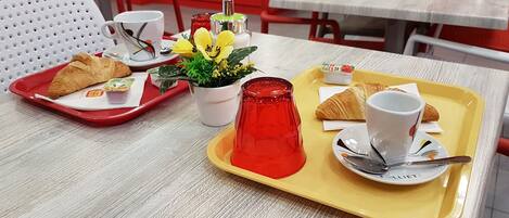 Una mesa de comedor con bandejas de comida, incluyendo croissants, un florero de vidrio rojo y una planta en maceta.