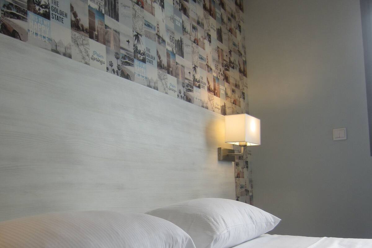 Una cama bien hecha con sábanas y almohadones blancos, una pared con un collage de imágenes, y una lámpara fijada a la pared.