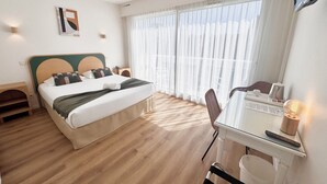 A hotel room with a bed, a desk, and a chair. There is a window with curtains, a wall-mounted light, and a picture on the wall. - Hotel Astoria (Saint-Etienne)