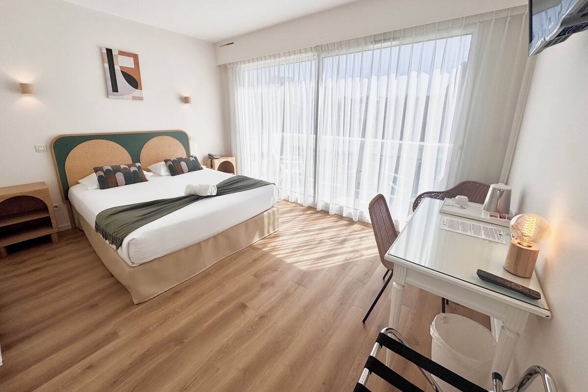 Habitación de hotel con cama, escritorio y silla. Se ve una ventana con cortinas, una lámpara de pared e un cuadro en la pared.