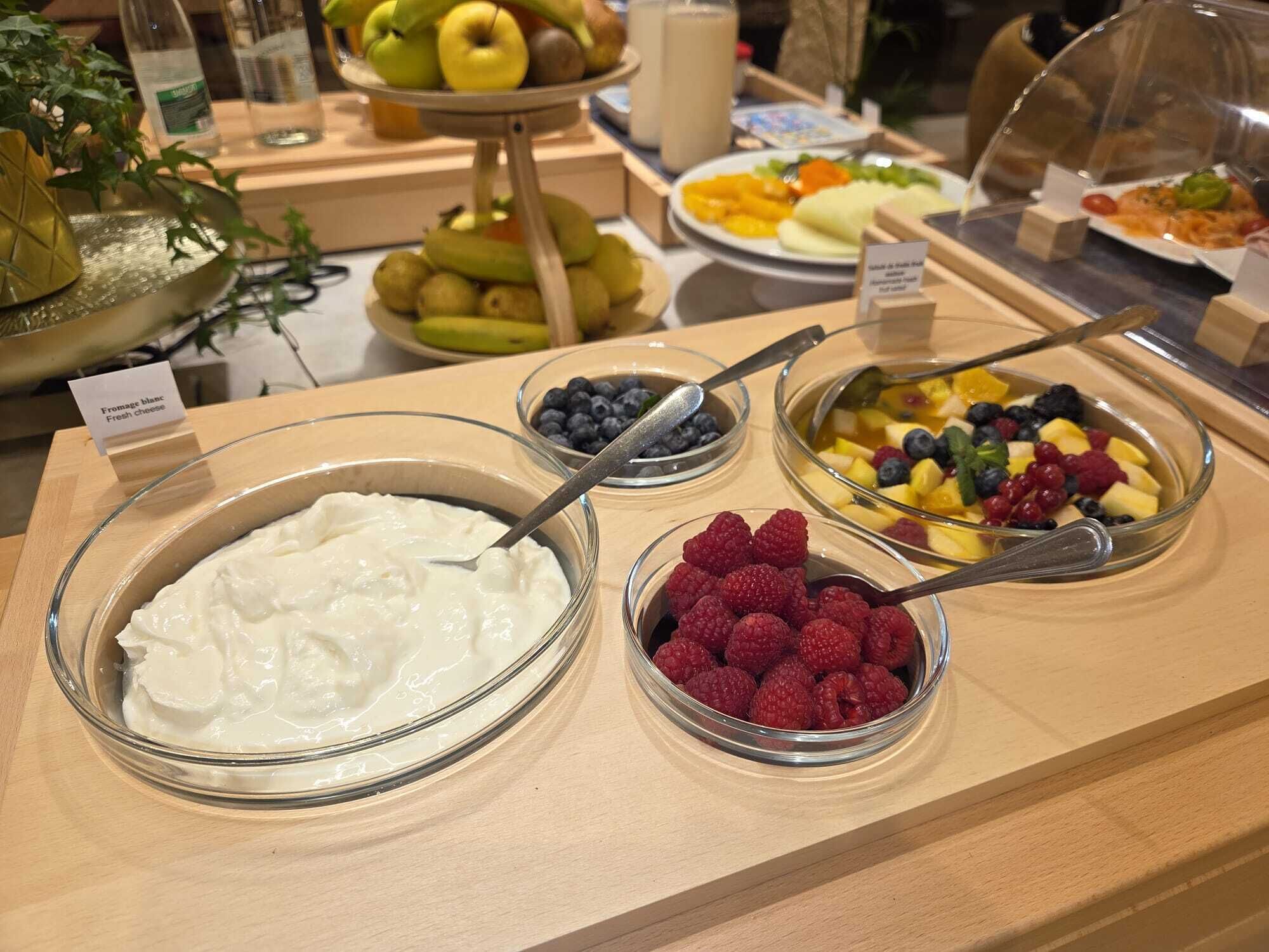 A buffet with various food items including yogurt, blueberries, raspberries, and a fruit salad.