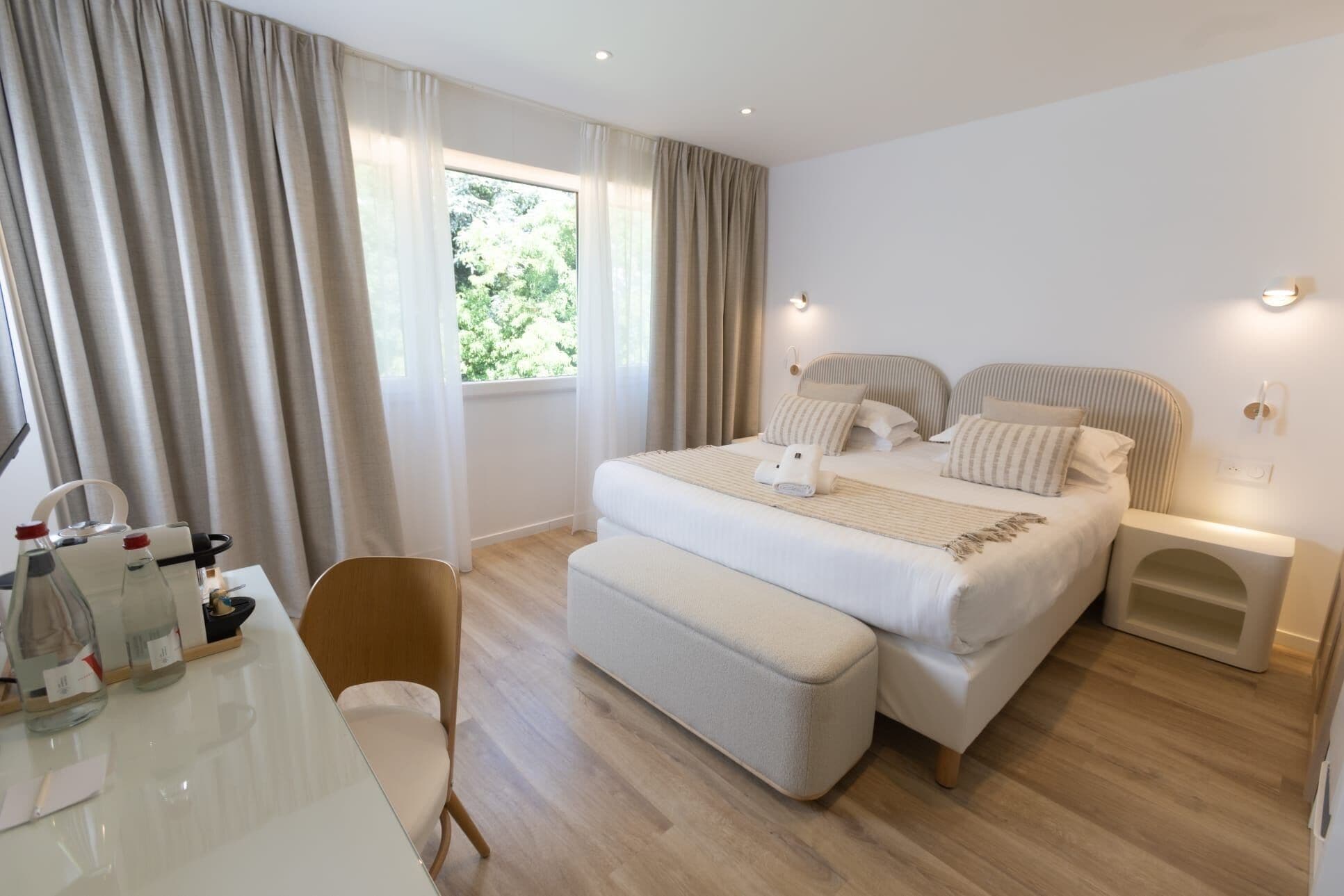 Habitación de hotel con una cama grande, mesitas de noche, una silla, un escritorio con artículos de tocador y una ventana con cortinas.