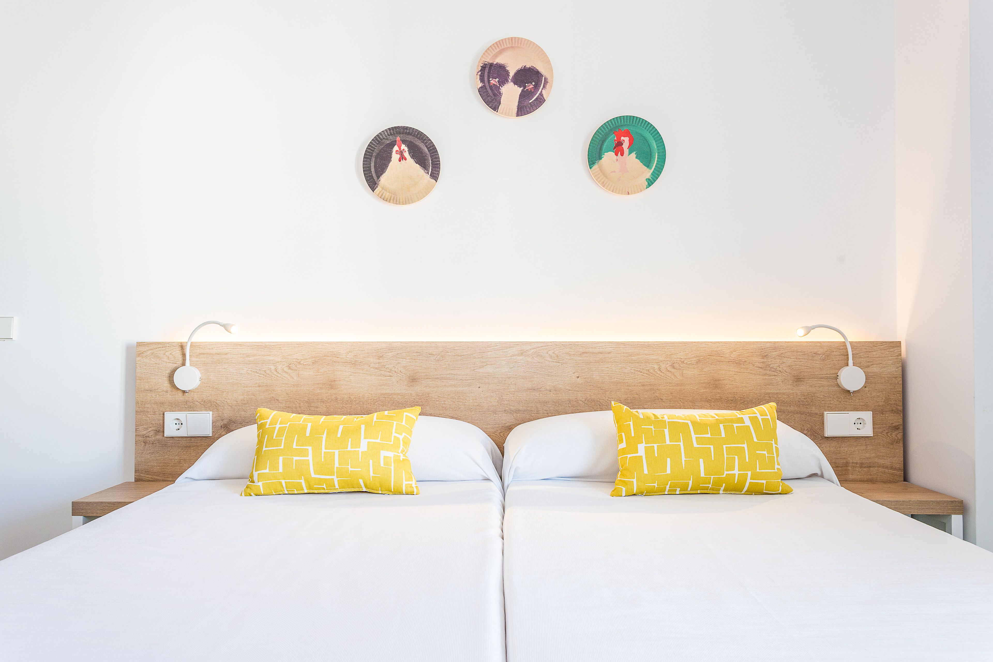 A double bed with white linens and two yellow patterned pillows, a wooden headboard, and a wall-mounted light fixture.