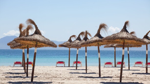 Una playa con sombrillas de paja y sillas rojas para tomar el sol.