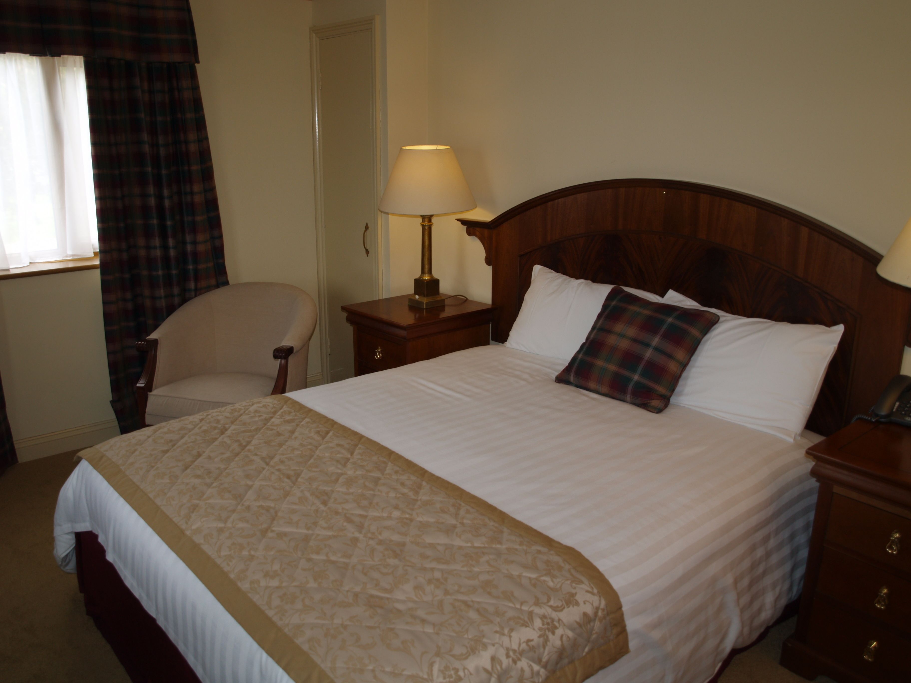 A neatly made bed with a plaid pillow, a bedside lamp, and a chair in a hotel room.