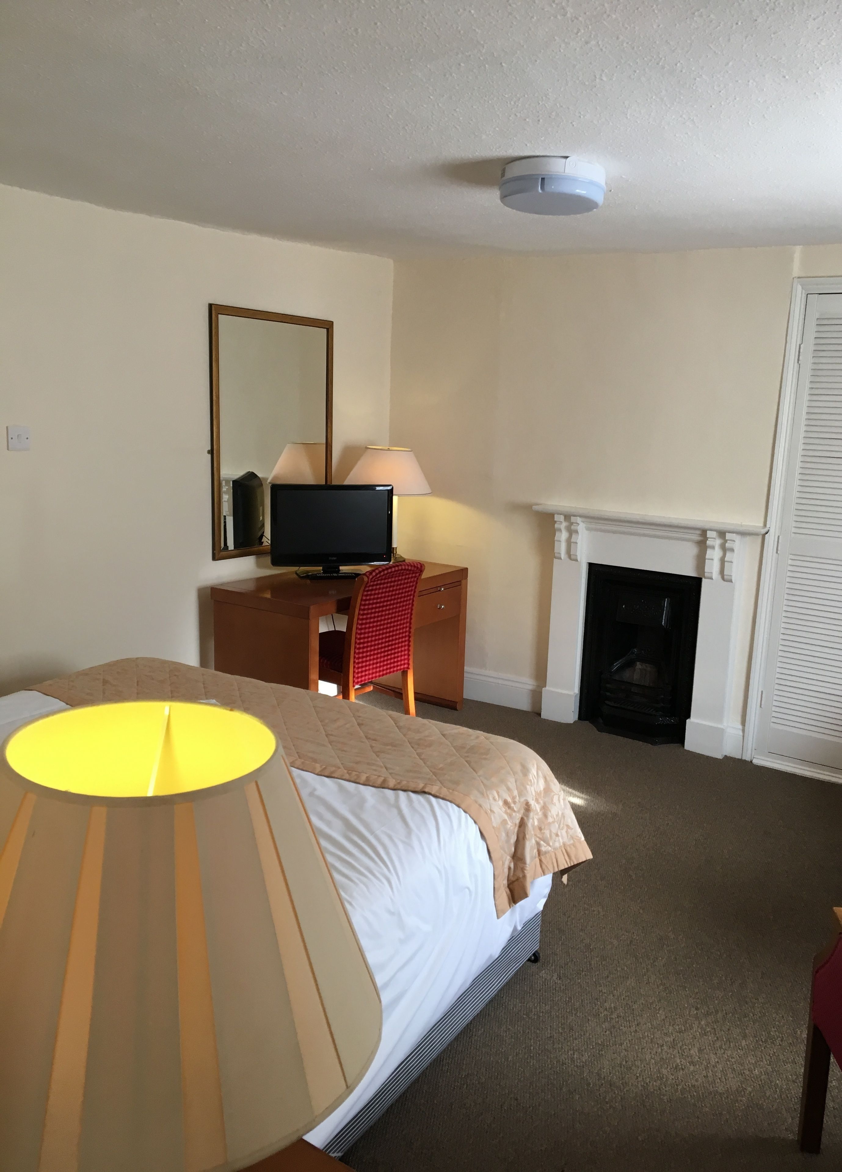 A hotel room with a bed, a desk with a computer, a chair, a lamp, a mirror, and a fireplace.