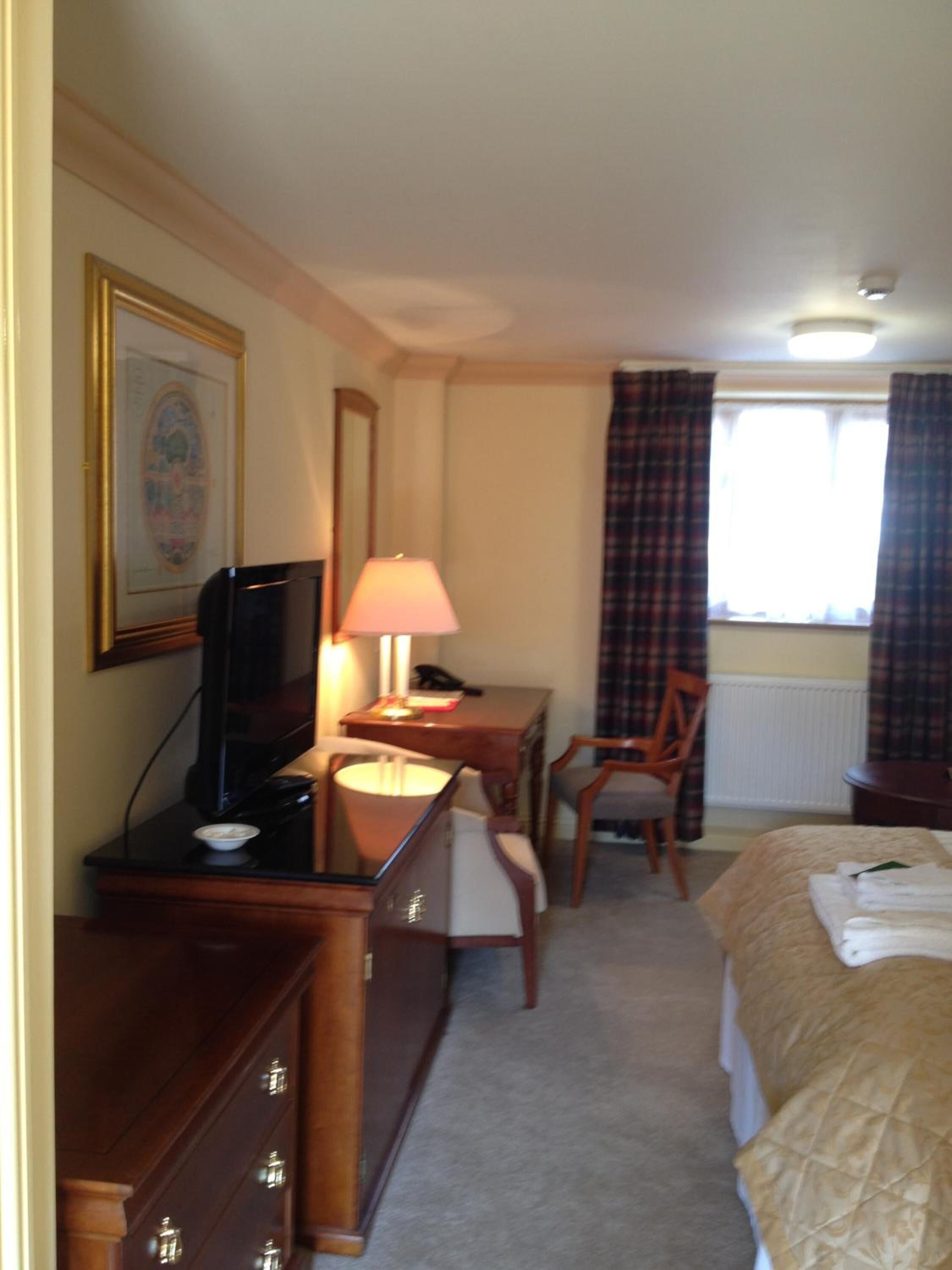 A hotel room with a bed, a desk with a lamp, a television, a chair, and a framed picture on the wall.