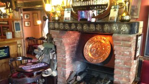 A cozy pub with a brick fireplace, wooden furniture, and a bar area with various bottles and glasses.