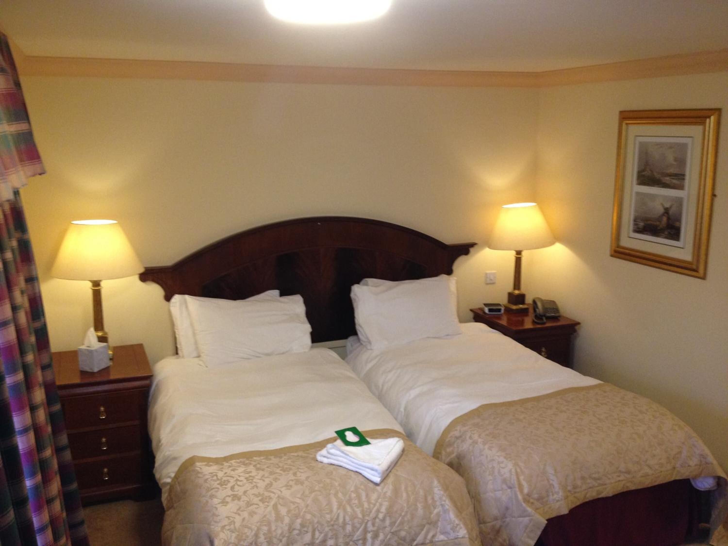 A hotel room with two beds, each with a wooden headboard, white linens, and a bedside table with a lamp and a telephone.