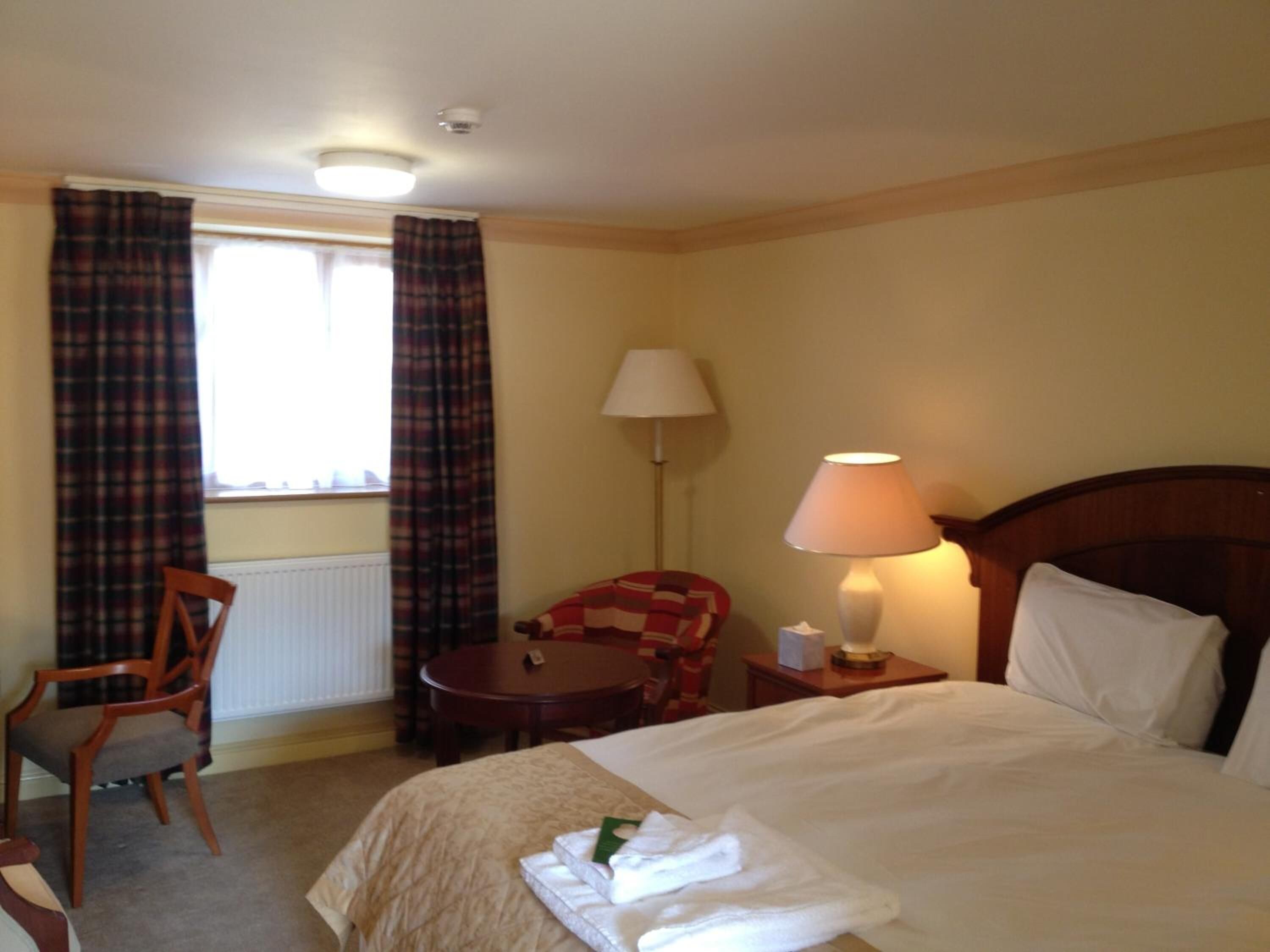 A hotel room with a bed, a chair, a table, a lamp, and curtains.
