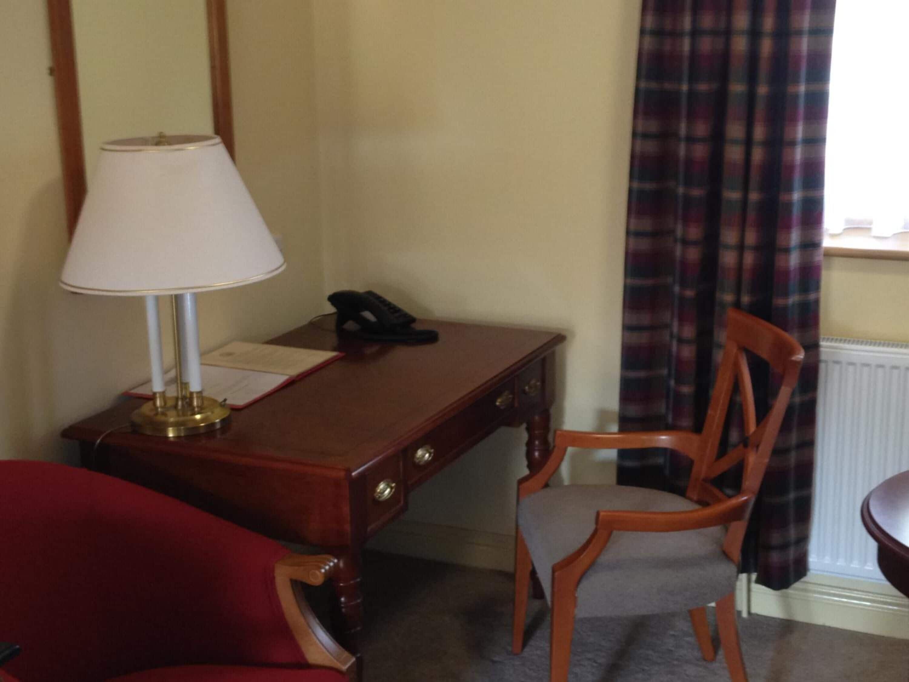 A wooden desk with a lamp, a telephone, and a chair in a room with a plaid curtain and a radiator.